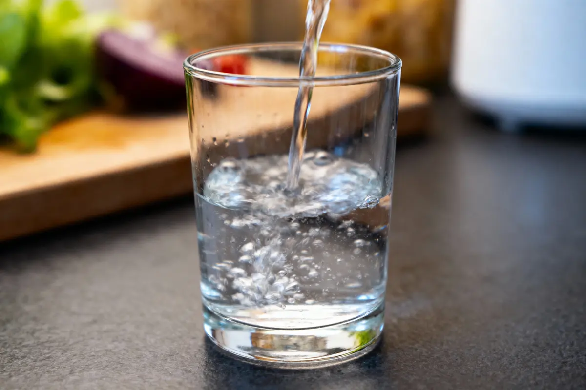 Gros plan d'un verre se remplissant d'eau pétillante, surface sombre, arrière-plan flou, aliments indistincts.