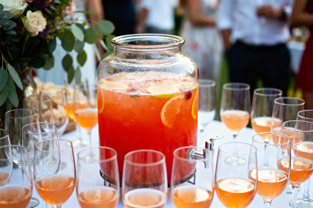 Un grand bocal de punch aux fruits avec verres autour, table blanche, décorations florales, invités flous en arrière-plan.