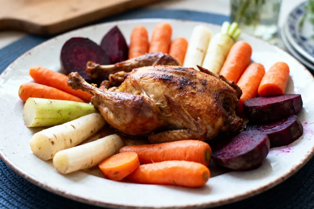 Poulet rôti entouré de légumes : carottes, betteraves, panais et poireaux, présenté sur une assiette blanche.