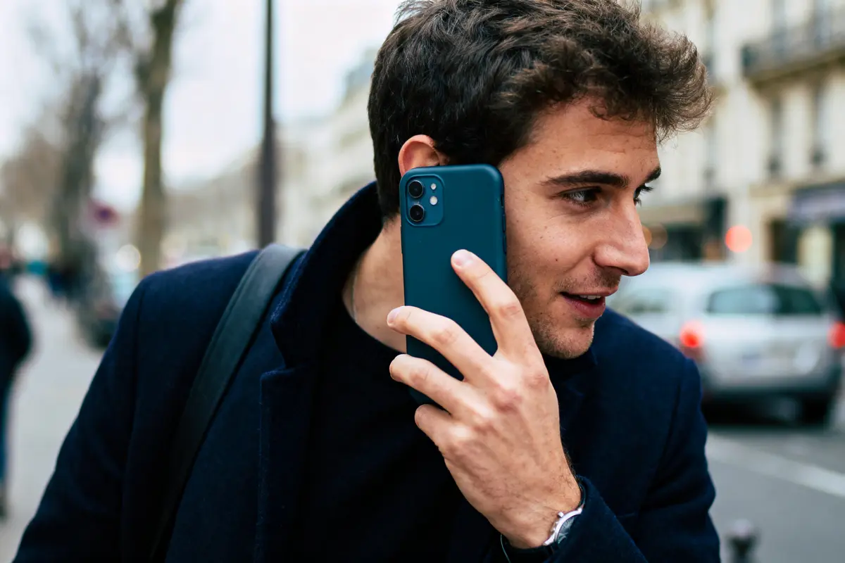 Homme parlant au téléphone dans la rue, portant un manteau sombre, arrière-plan urbain flou.