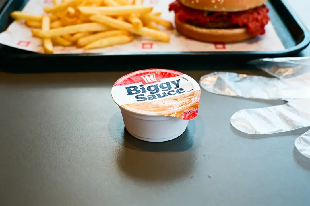 Sauce "Biggy" sur table, avec frites, burger et gants en fond.