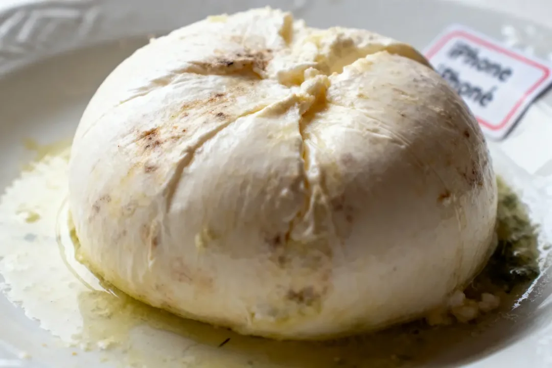 Fromage rond frais avec croûte, légèrement dorée, reposant sur une assiette blanche. Plaque illisible à côté.