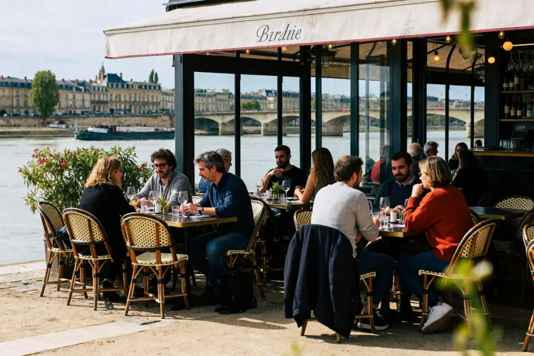 Des personnes dînent en terrasse au bord de l'eau, avec vue sur un pont et des bâtiments historiques.