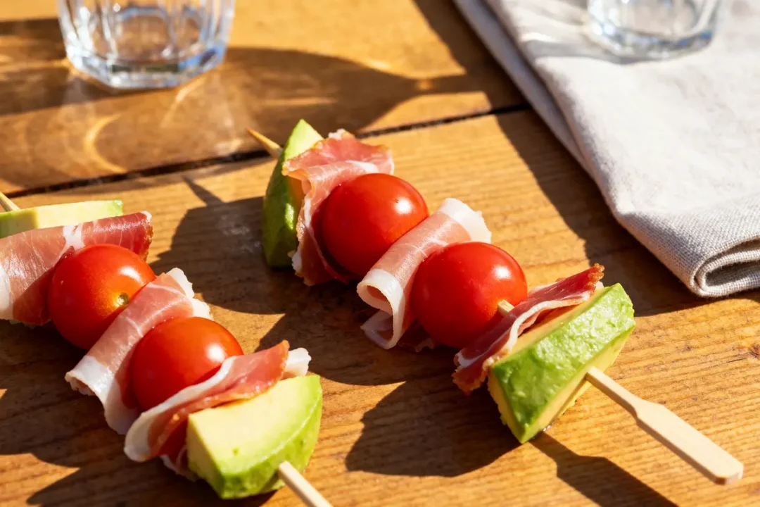 Brochettes apéro avec avocat, jambon et tomate sur table en bois ensoleillée
